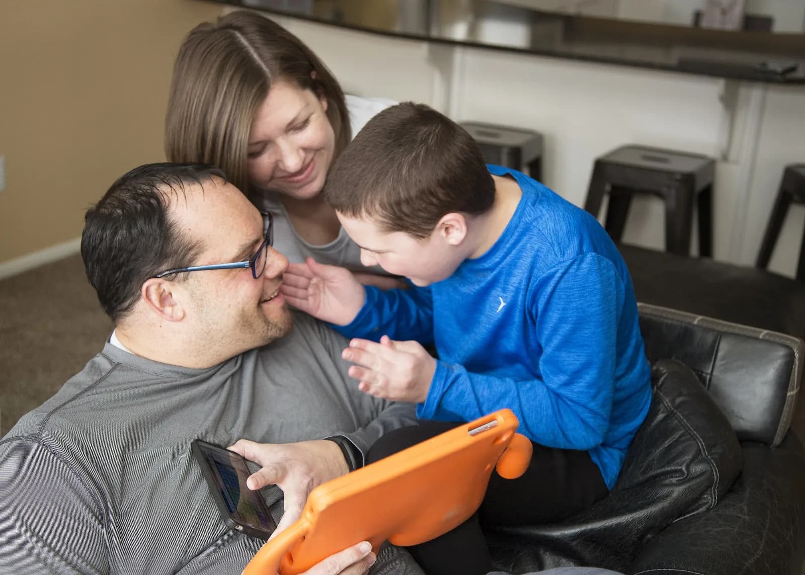 Two parents and a child sitting together on a sofa, looking at a tablet running a communication app